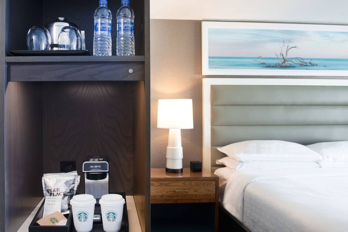 A hotel room with a bed, a nightstand with a lamp, a tray with coffee cups and a coffee machine, a shelf with water bottles and glasses, and a painting above the bed.
