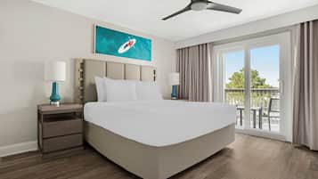 A hotel room with a large bed, two bedside tables with lamps, a ceiling fan, a painting on the wall, and a view of the outdoors through a sliding glass door.