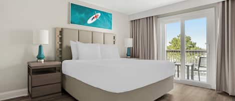 A hotel room with a large bed, two bedside tables with lamps, a ceiling fan, a painting on the wall, and a view of the outdoors through a sliding glass door.