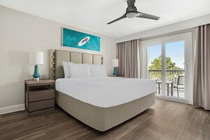 Quarto de hotel com uma cama grande, duas mesas de cabeceira com abajures, um ventilador de teto, um quadro na parede e vista para o exterior por uma porta de vidro deslizante.