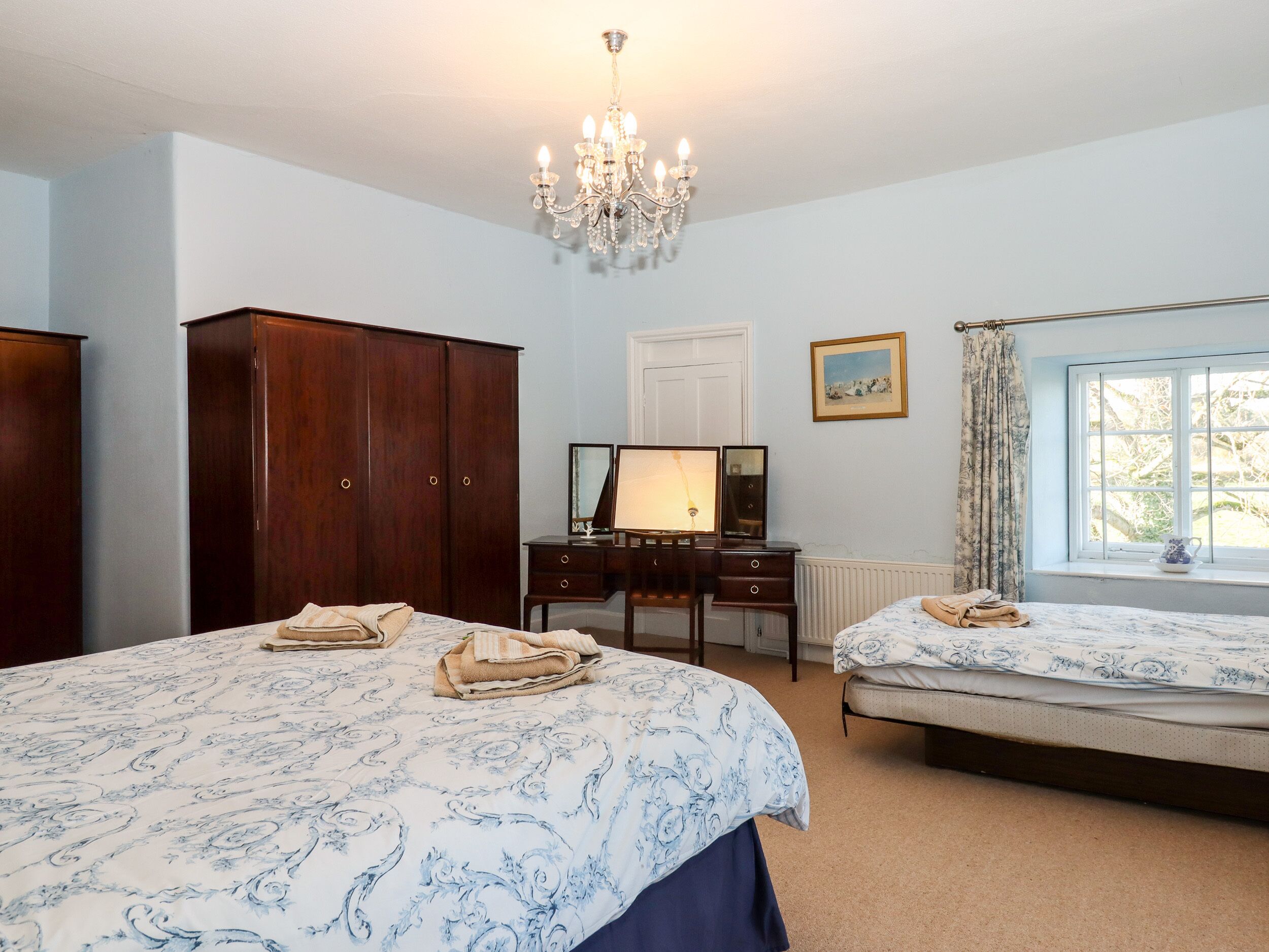 A bedroom with two beds, a dresser, a mirror, a chandelier, and a window with curtains.