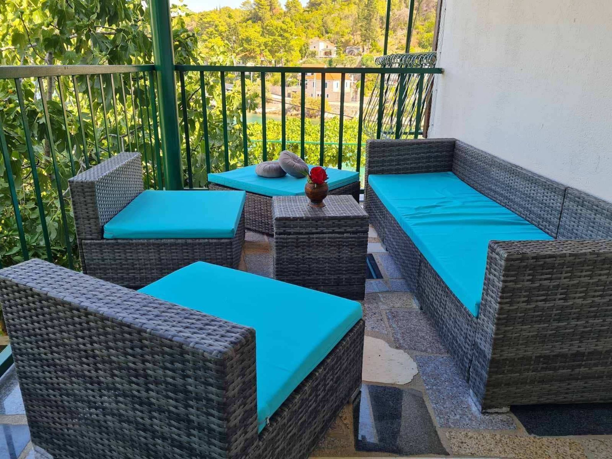 Un balcón con muebles de mimbre y un cojín azul, con vistas a un jardín exuberante y edificios.