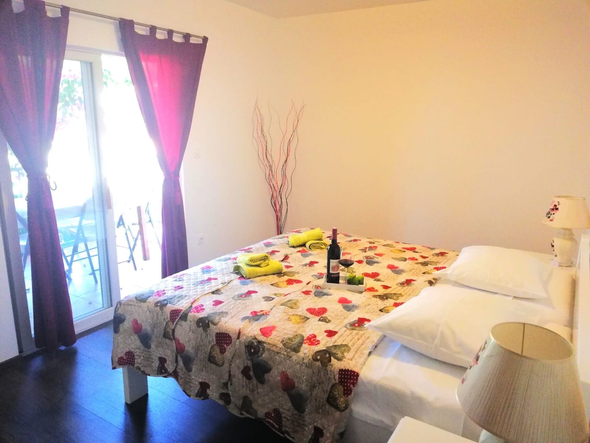 Habitación de hotel con cama, ventana con cortinas, mesita de noche con lámpara y una pequeña mesa con una botella y vasos.