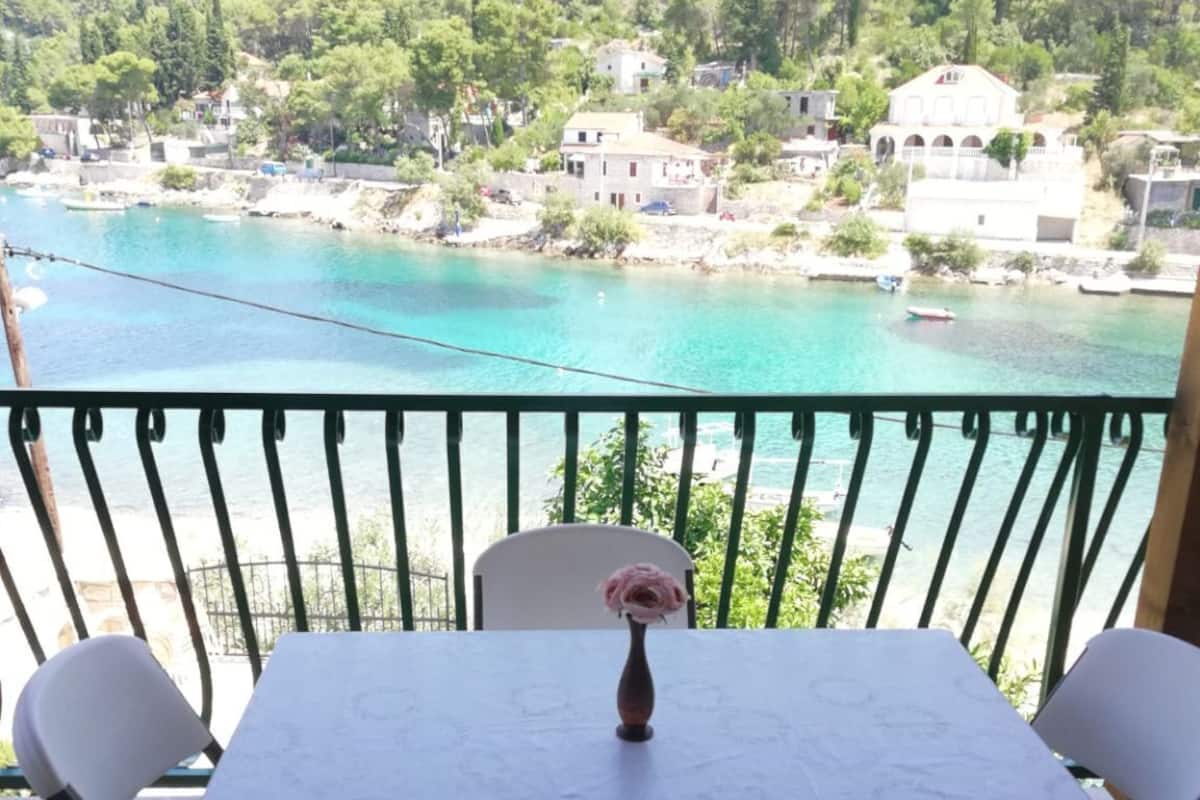 Ein Balkon mit Tisch und Stühlen mit Blick auf ein türkisblaues Gewässer und eine ansteigende Hanglage mit Gebäuden.
