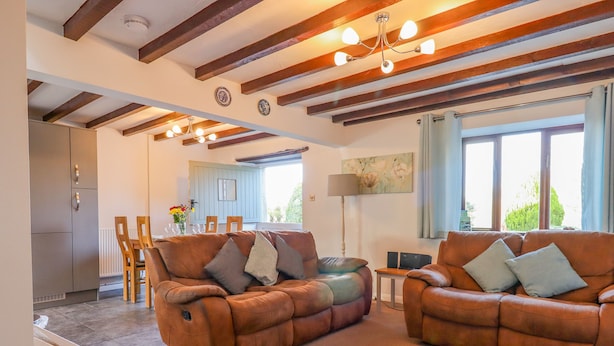 A spacious living room with two brown leather sofas, a dining area with wooden chairs, and a kitchen with a built-in refrigerator. - Oak Leaves Cottage (Newton Abbot)