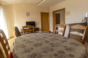 A room with a round table covered in a floral tablecloth, wooden chairs, a television on a stand, a clock on the wall, and a kitchen area visible through an open door. - Apartments Marija (Makarska)