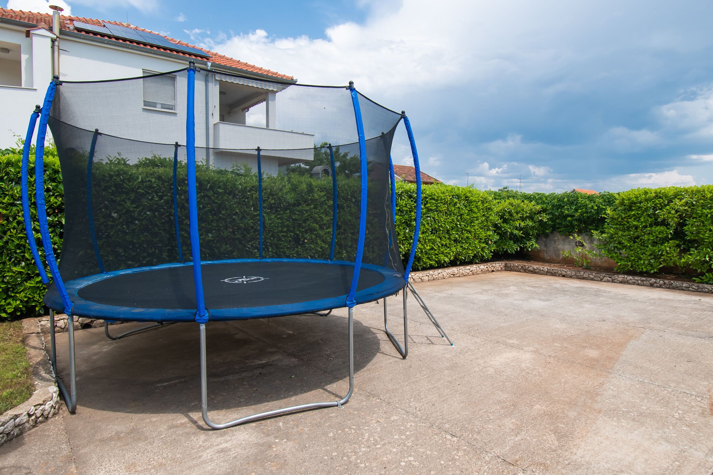 A trampoline with a safety net, set on a concrete patio, surrounded by greenery and a house in the background.