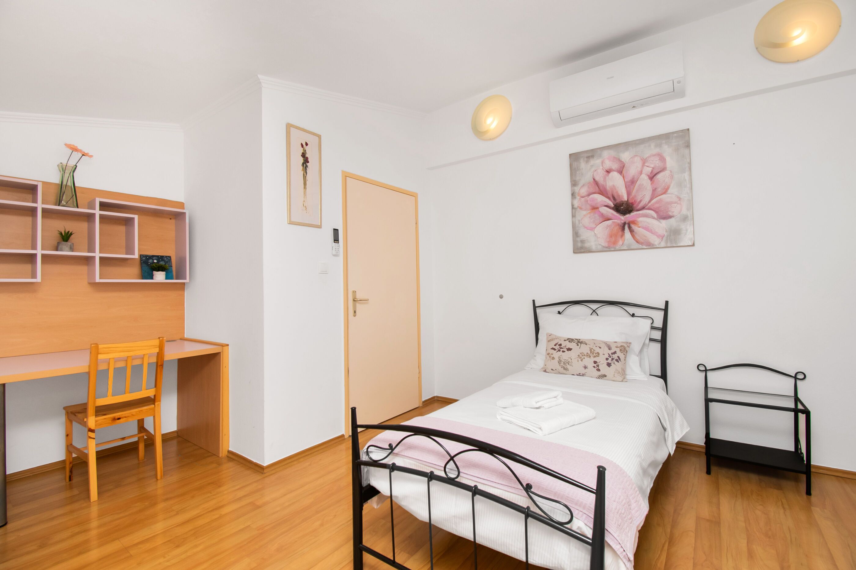 A bedroom with a bed, a desk, a chair, a shelf with decorative items, and a door.