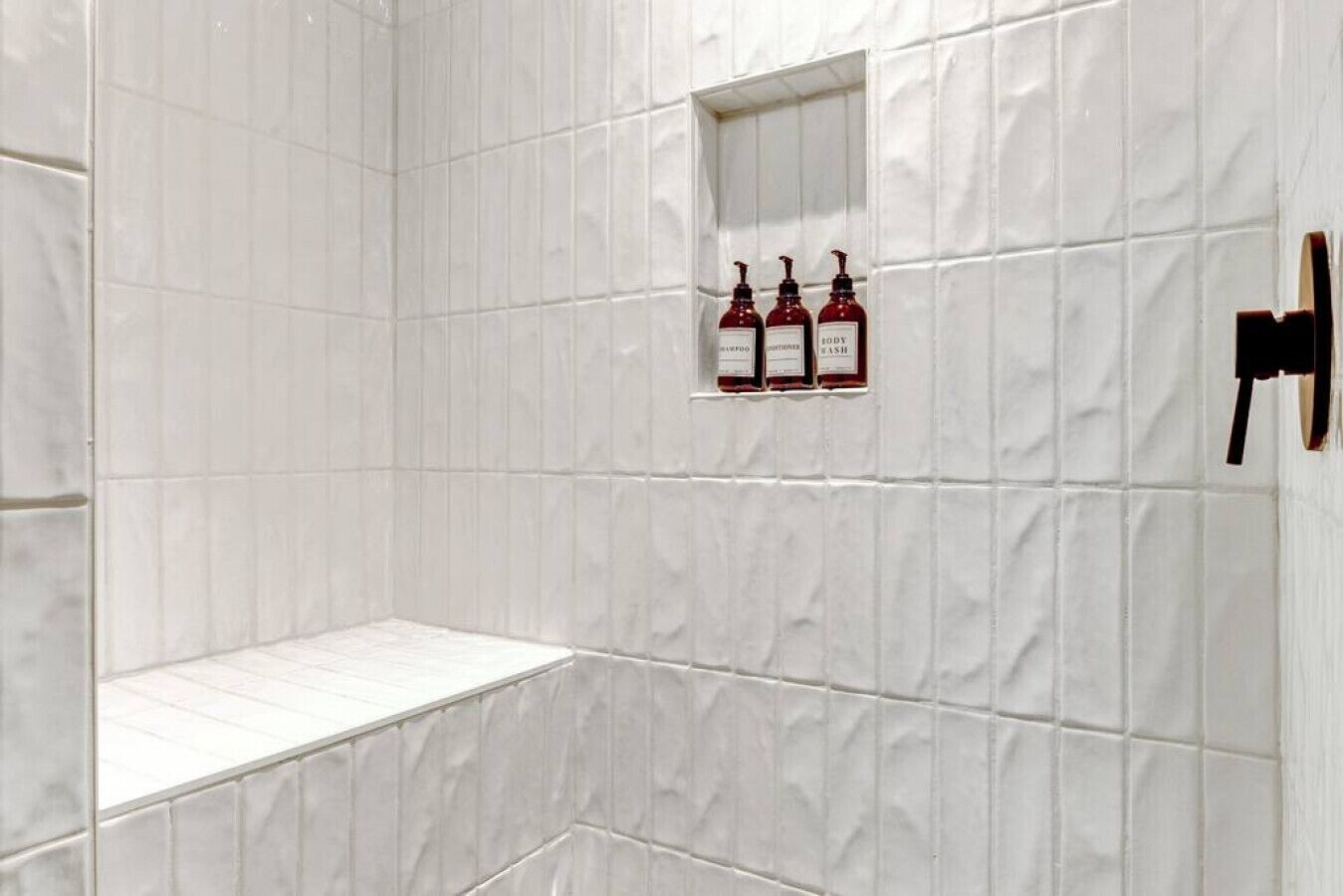A bathroom with white tiled walls, a small shelf, and three bottles of shampoo and conditioner.