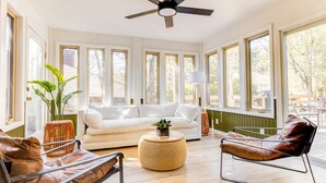 A spacious living room with a white sofa, leather chairs, and a round coffee table. Large windows provide ample natural light and views of the outdoors.