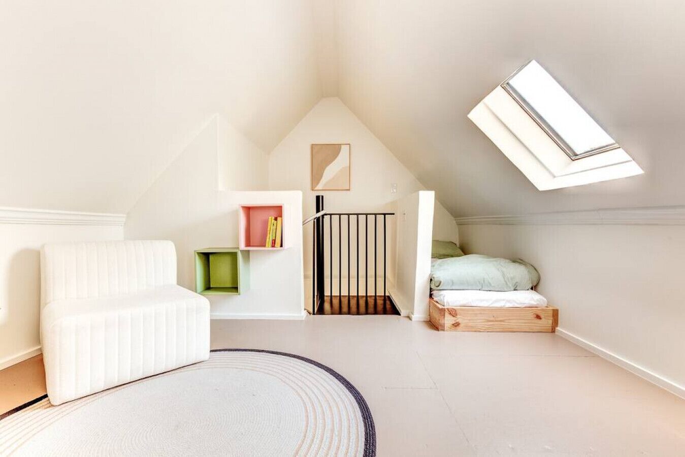 Une chambre mansardée chaleureuse avec un plafond en pente, une lucarne, un canapé blanc, un petit lit et une bibliothèque verte et rose.