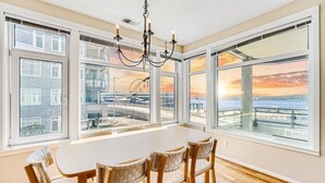 Un espace repas comprenant une grande table et des chaises, de grandes fenêtres offrant une vue sur le bord de l’eau et sur le coucher de soleil, ainsi qu’un lustre.