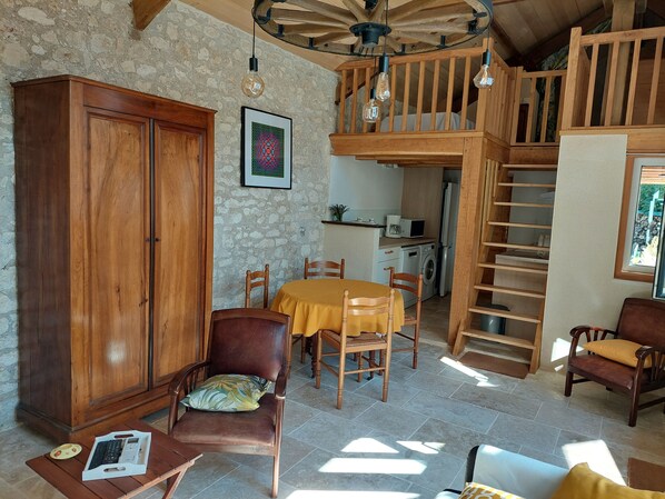 A cozy living room with a wooden cabinet, a dining table with chairs, a staircase, and a kitchen area in the background.