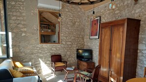 Ein gemütliches Wohnzimmer mit Steinwand, einer Holzkommode, einem Fernseher auf einem Holzboden, einer Couch mit gemusterten Kissen und einem Esstisch mit gelber Tischdecke.