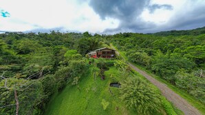 Une maison en bois isolée, de plain-pied, entourée d’une forêt tropicale dense, et accessible par un sentier de terre.