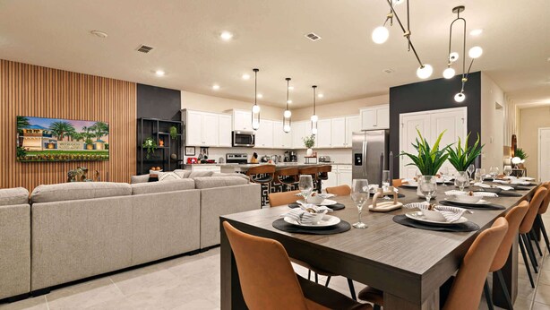 A modern kitchen with white cabinets, a dining area with a long table set for six, and a living area with a sofa and a TV mounted on the wall.