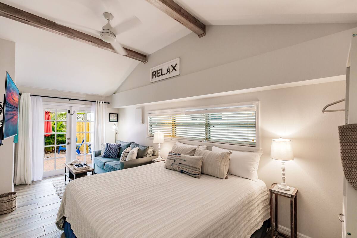 A bedroom with a large bed, a bedside table with a lamp, a window with blinds, a ceiling fan, and a sign that says "RELAX".