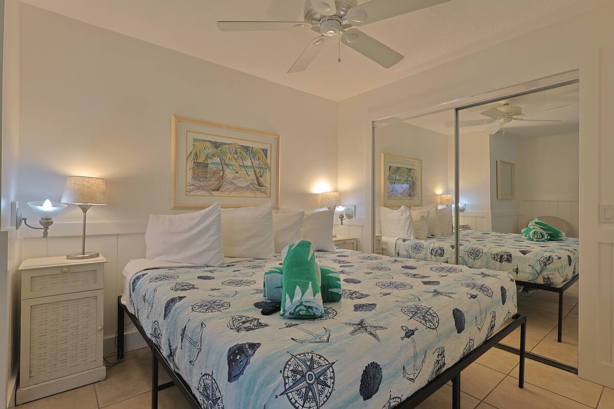 A bedroom with a bed, a nightstand, a lamp, a ceiling fan, and a painting on the wall.