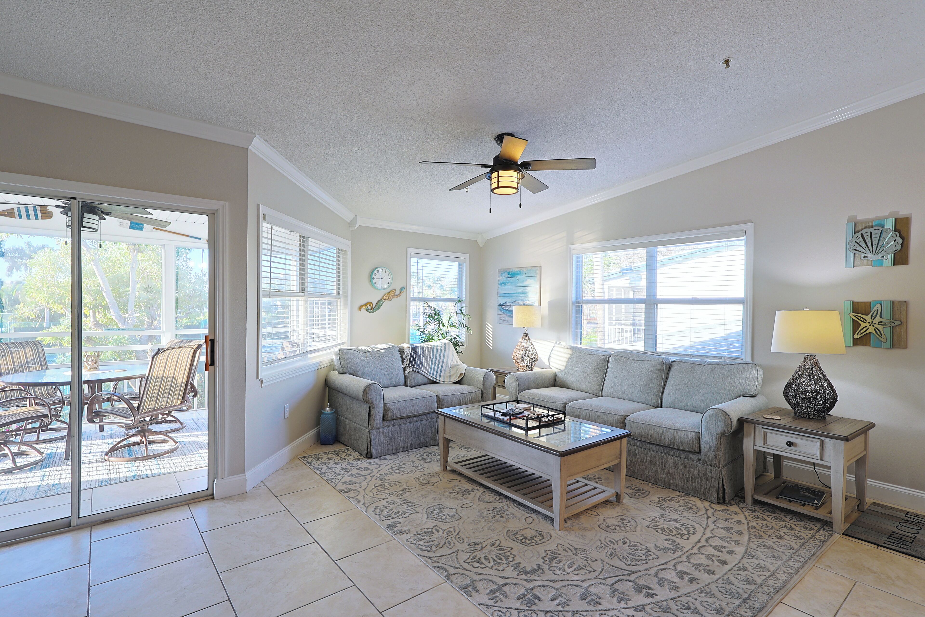A living room with two sofas, a coffee table, and a ceiling fan. There are windows and a door leading to an outdoor patio.