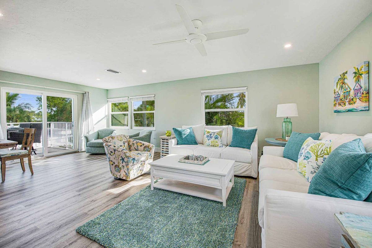 A spacious living room with a white sofa, a floral armchair, and a coffee table. There is a ceiling fan, a painting on the wall, and a view of the outdoors through large windows.