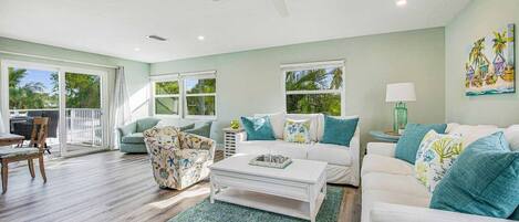 A spacious living room with a white sofa, a floral armchair, and a coffee table. There is a ceiling fan, a painting on the wall, and a view of the outdoors through large windows.