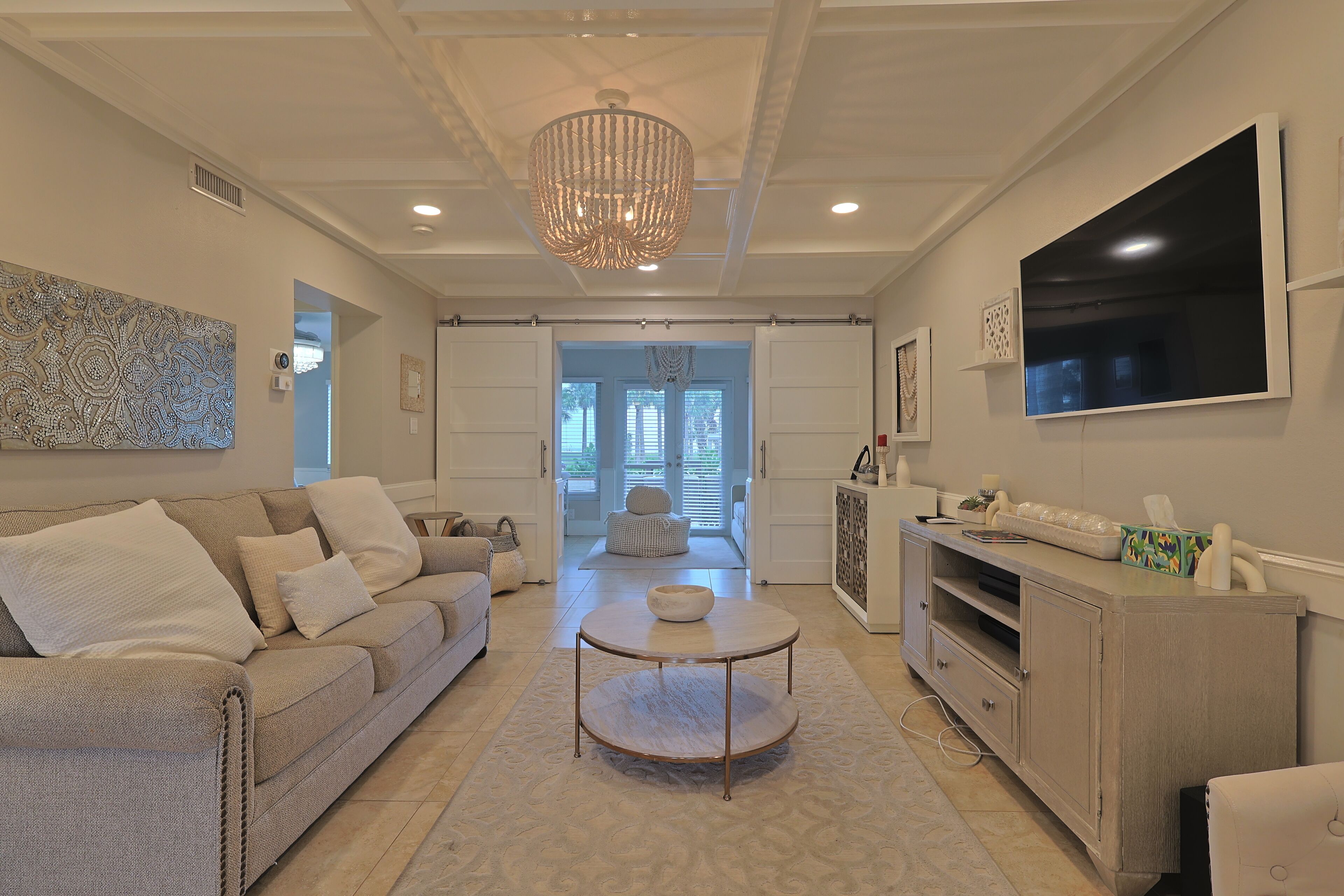 A modern living room with a beige sofa, a round coffee table, a TV mounted on the wall, and a chandelier.