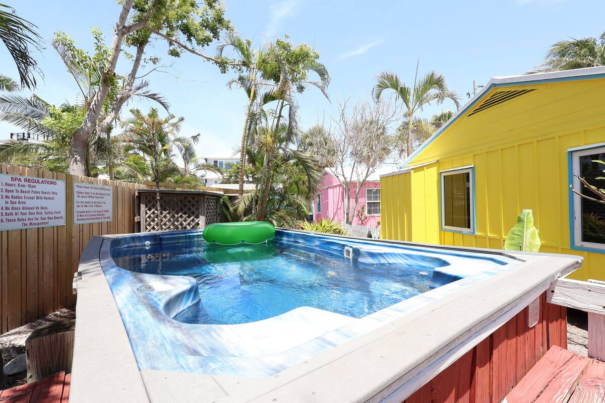 A small outdoor hot tub area with a wooden deck, surrounded by palm trees and a yellow building.