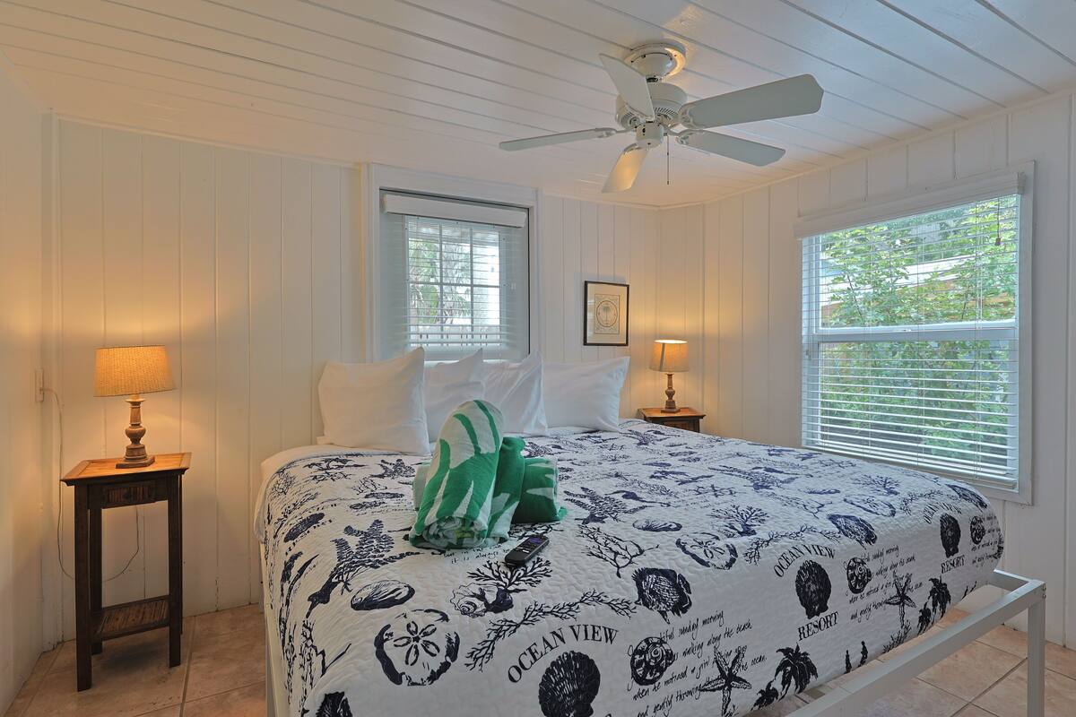 A bedroom with a bed, two bedside lamps, a ceiling fan, and a window with blinds.