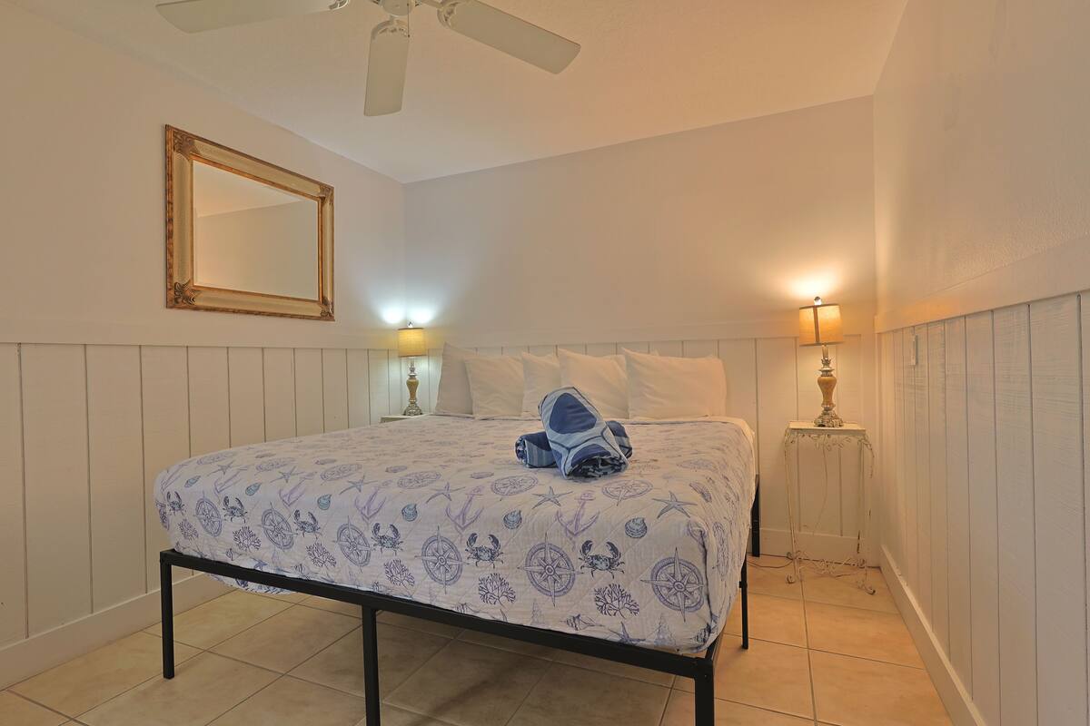 A bedroom with a bed, two bedside lamps, a ceiling fan, and a mirror on the wall.