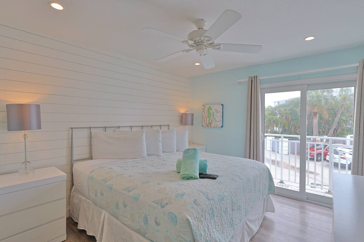 A bedroom with a bed, a ceiling fan, a nightstand, a lamp, and a window with a view of the outside.