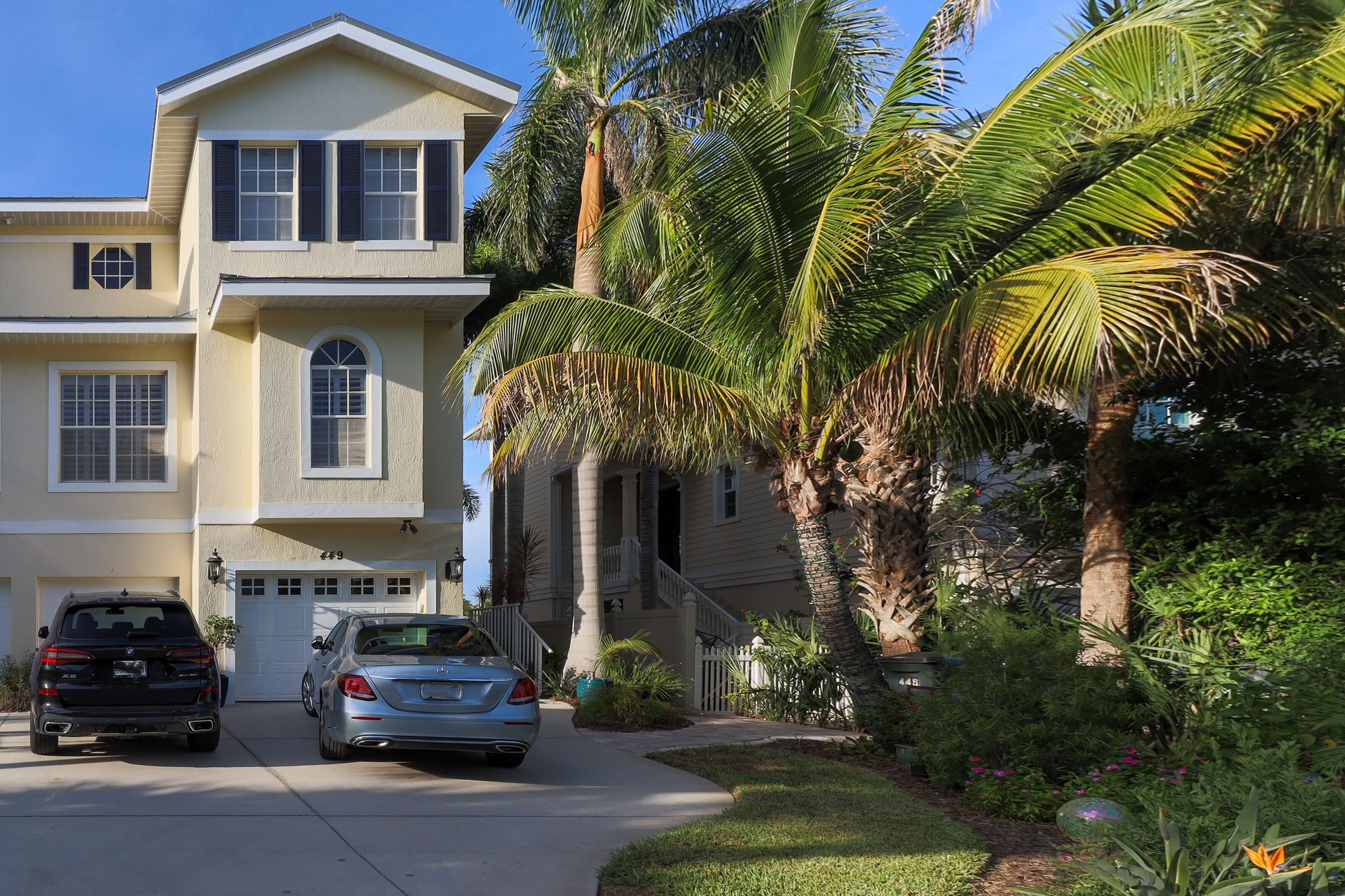 A two-story house with a garage, parked cars, and a well-maintained garden with palm trees.