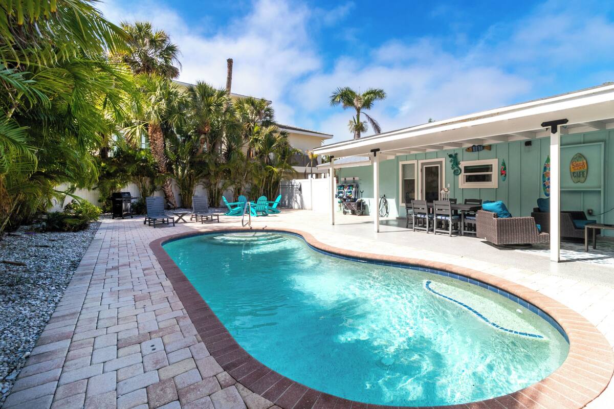 A swimming pool with a brick border, surrounded by palm trees and a covered patio area with outdoor furniture.