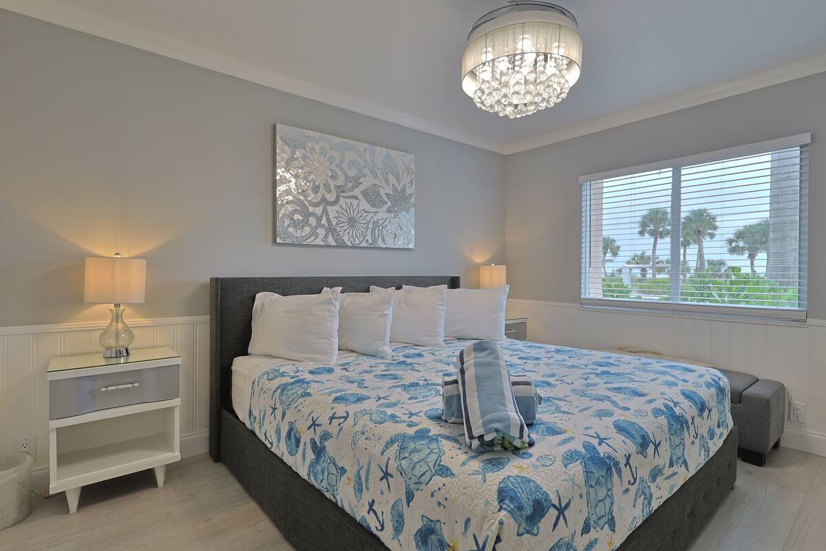 A bedroom with a large bed, a nightstand, a chandelier, a window with blinds, and a decorative wall piece.