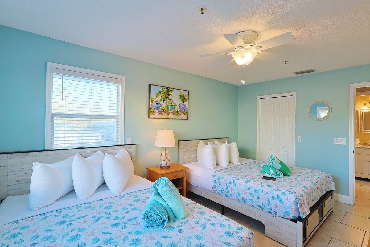 A bedroom with two beds, a ceiling fan, a window with blinds, a painting, a nightstand with a lamp, and a door leading to another room.