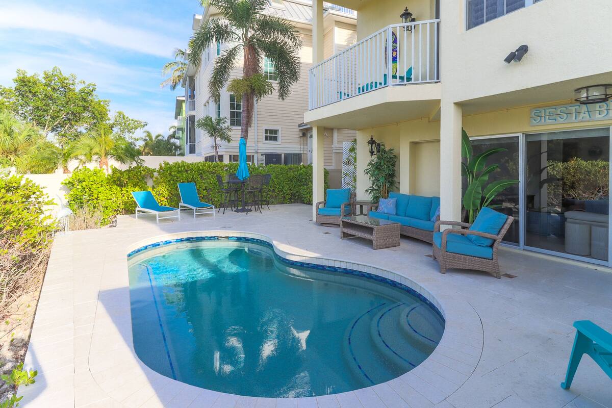A pool area with lounge chairs and a small table, surrounded by greenery and a building with a balcony.