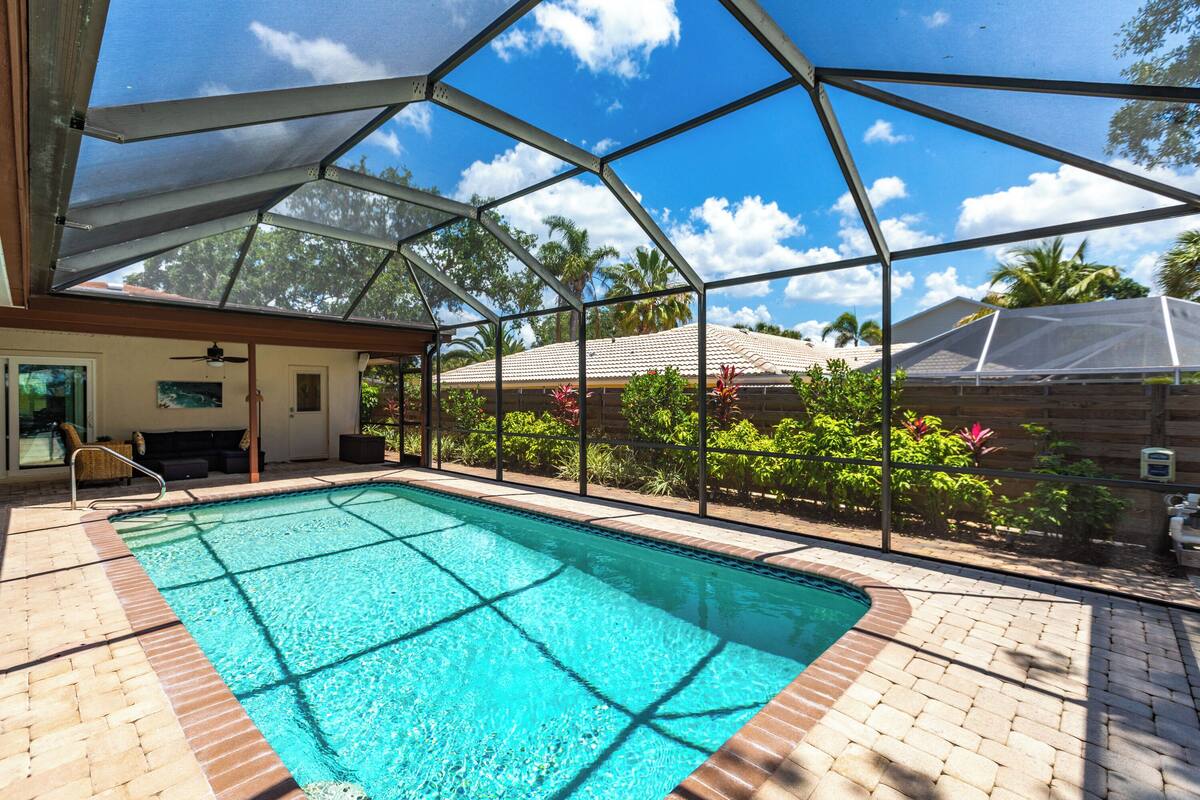 A swimming pool with a covered area, surrounded by a brick patio and lush greenery.