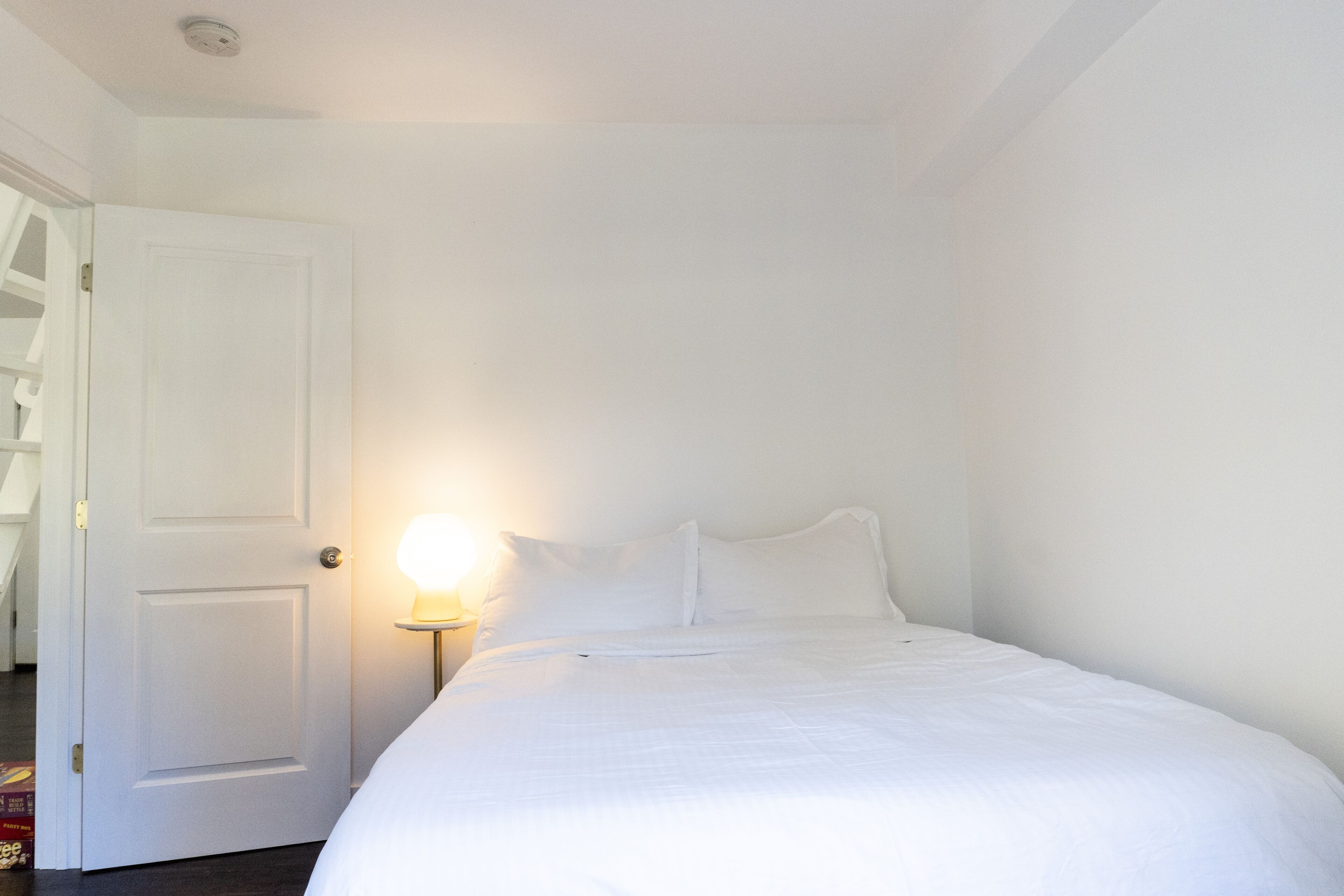 Una habitació blanca amb un llit, una porta i una tauleta de nit amb una làmpada.