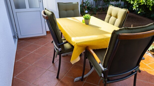 Un patio exterior con una mesa amarilla y cuatro sillas, una planta en maceta y una puerta.