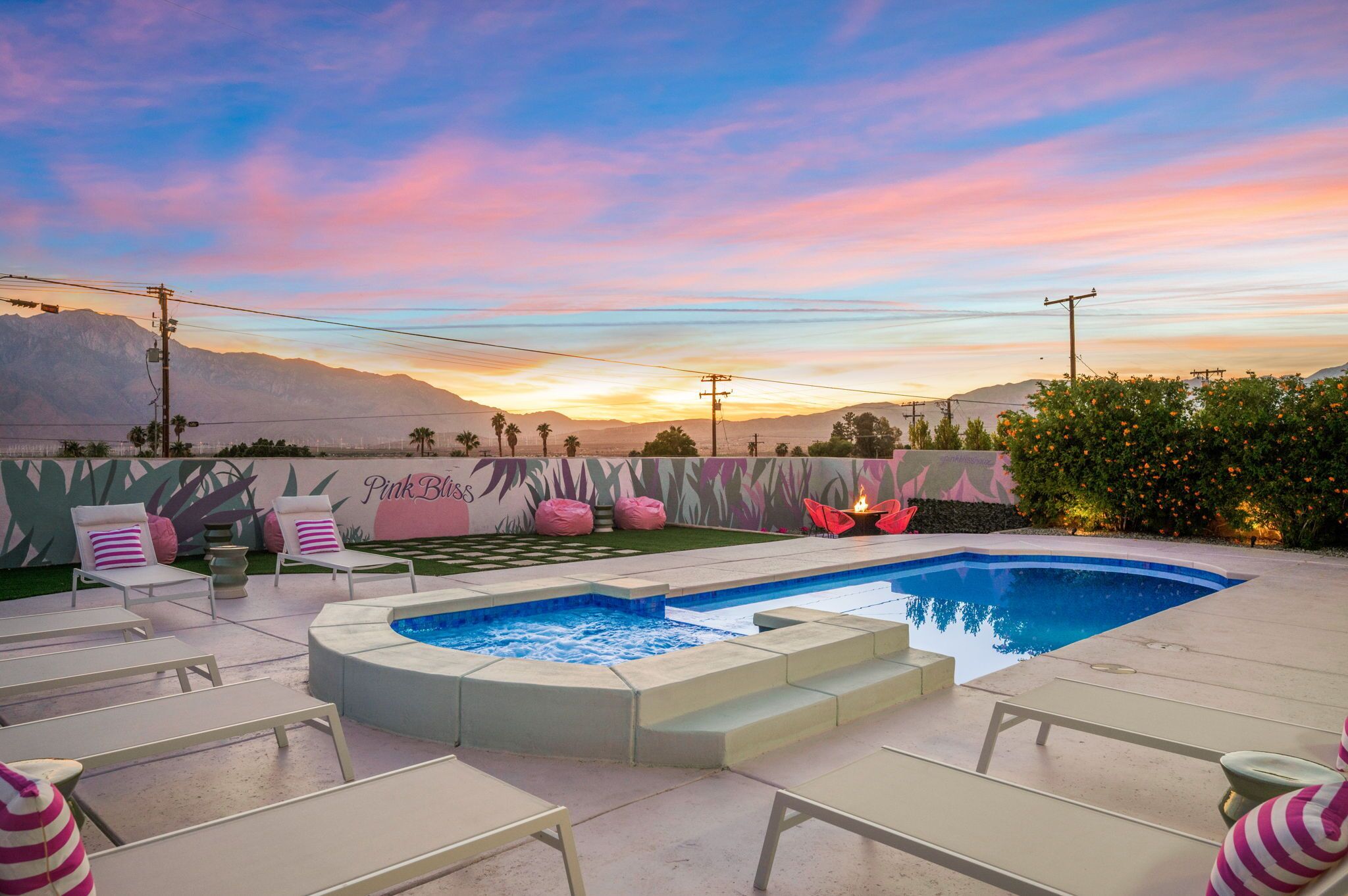 A pool area with lounge chairs and a hot tub, surrounded by a decorative wall with a 'Pink Bliss' mural, set against a sunset sky.