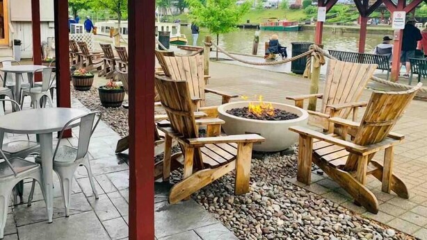 An outdoor seating area with wooden Adirondack chairs and a fire pit, surrounded by a stone pathway and a view of a canal with boats. - *Posh 2BR in the Heart of the Pittsford Village* (Pittsford)