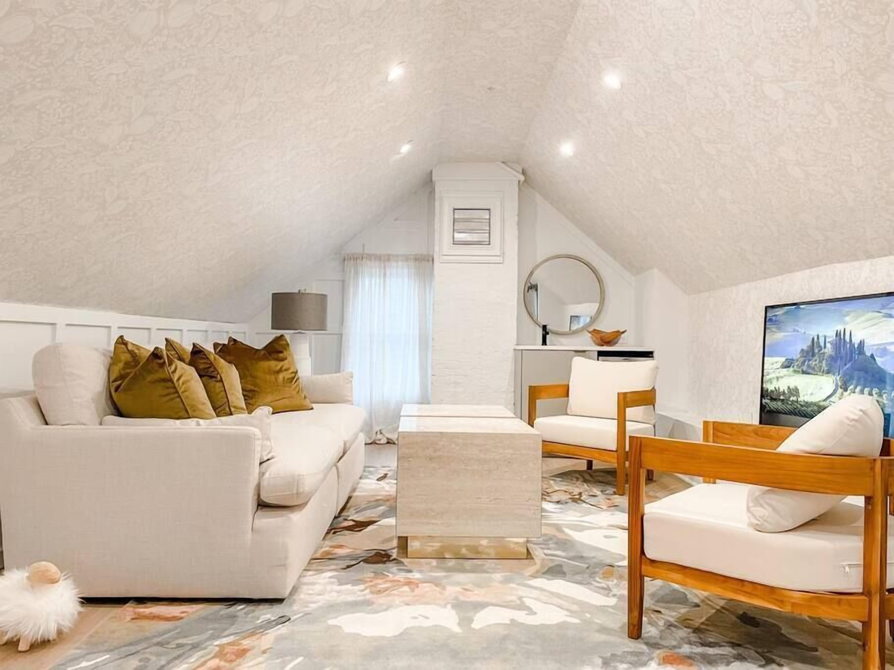 An attic bedroom with a white ceiling, a white sofa with mustard pillows, a wooden chair, a small table, and a large TV on the wall.