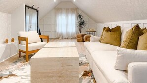 A spacious attic bedroom with a high ceiling, a large window with sheer curtains, a wooden chair, a white sofa, and a small table with a plant. - *Posh 2BR in the Heart of the Pittsford Village* (Pittsford)