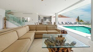 A modern living room with a glass coffee table, beige sofa, and a view of a swimming pool and mountains.