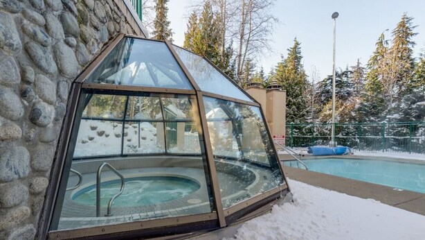 A glass-enclosed hot tub adjacent to a swimming pool, with a stone wall and snow-covered ground. - The Maple | Ski-in Ski-out 1 Bed w/ Hot Tub, Pool, Sauna, and Fireplace (Whistler)