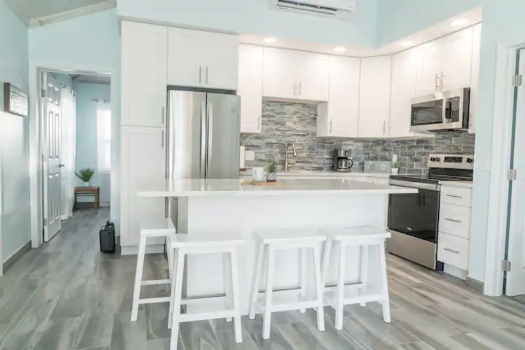 A modern kitchen with white cabinets, a central island with bar stools, stainless steel appliances, and a stone backsplash.