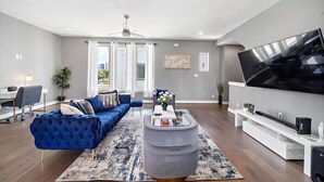 A modern living room with a blue sofa, a grey ottoman, a flat-screen TV, and a white entertainment unit. - Your lavish contemporary duplex | downtown Houston| Rooftop. (Houston)