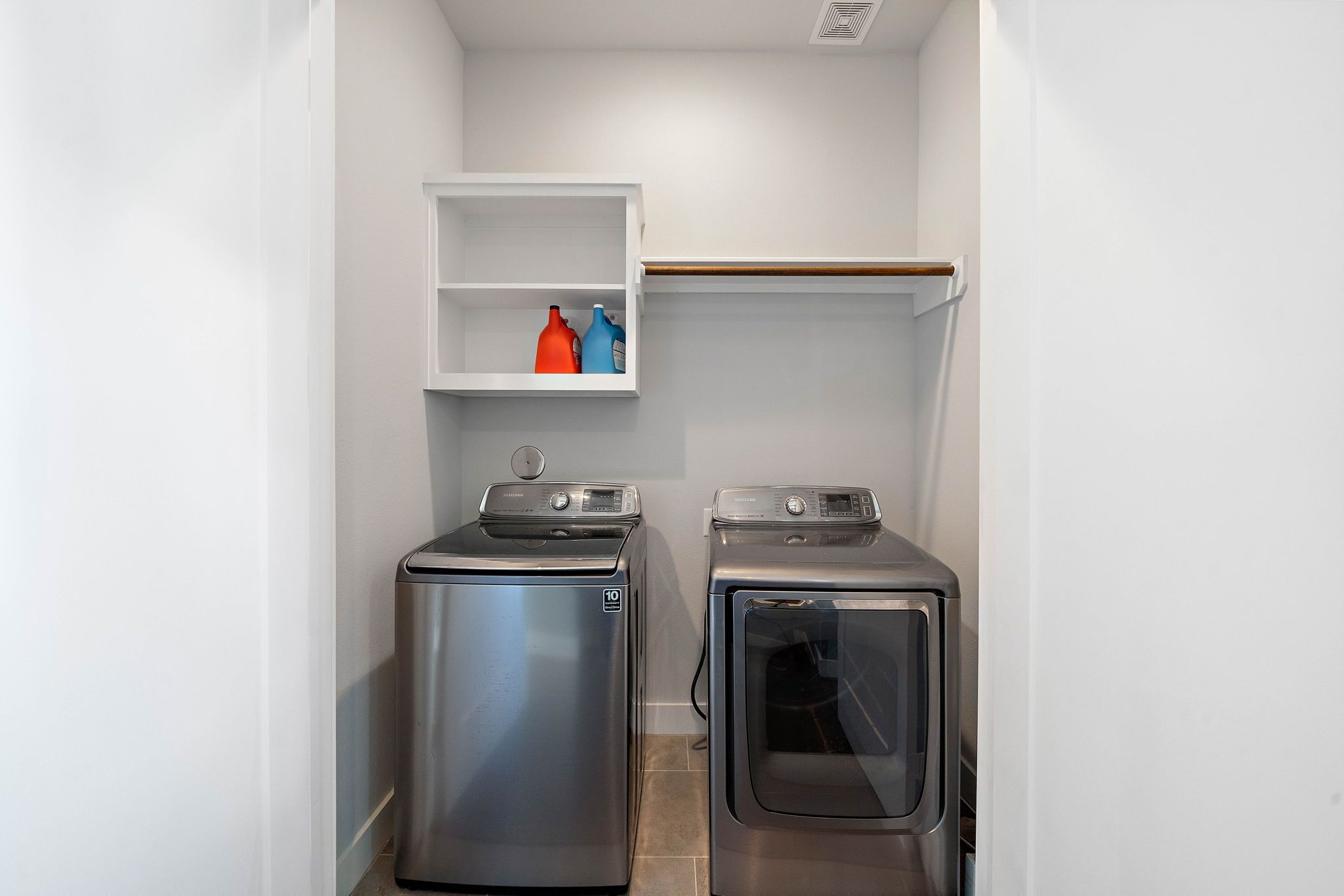 A laundry room with two stainless steel washers and dryers, a white shelf with cleaning supplies, and a wooden shelf above.