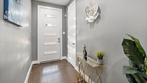 A hallway with a white door, a decorative wall piece, a small table with a plant, and a large green plant to the right. - Your lavish contemporary duplex | downtown Houston| Rooftop. (Houston)