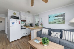 A compact living space with a kitchenette, a grey sofa with striped cushions, and a painting of a coastal landscape.