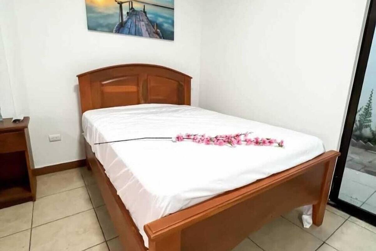 A bedroom with a wooden bed, a nightstand, a painting on the wall, and a view through a glass door.